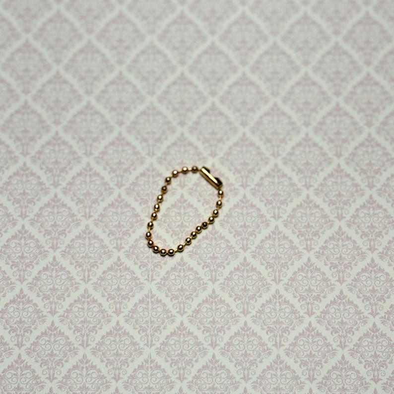 May include: A small gold-toned ball chain connector, also known as a bead chain connector, featuring a series of tiny spherical metal beads linked together in a circular loop with a cylindrical clasp mechanism to secure the ends.