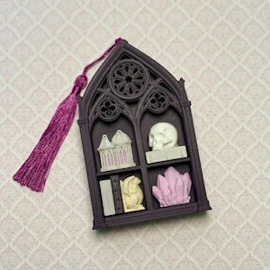 May include: A dark purple gothic window-shaped frame featuring a long magenta tassel. The frame contains four small decorative items: a white birdcage, a white skull on a book, a small book next to a dragon figurine, and a cluster of light pink crystals.