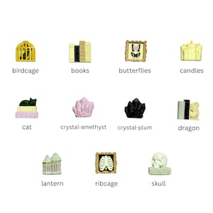 May include: A collection of eleven miniature decorative figurines including a yellow birdcage, books, framed butterflies, candles, a cat on books, amethyst and plum crystals, a dragon, a lantern, a framed ribcage, and a skull on a book.