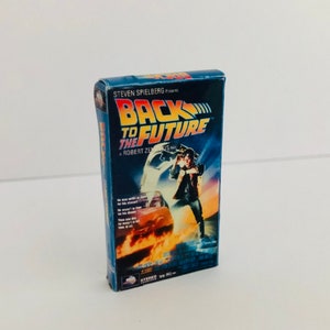 Puede incluir: Una cinta VHS en miniatura de la película "Regreso al Futuro". La portada muestra el título de la película en amarillo y azul, con una imagen de una persona frente a un coche en llamas. También se ve el texto "Steven Spielberg Presents".