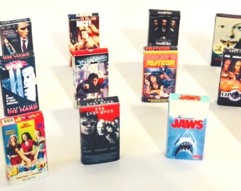 May include: A collection of VHS tapes featuring popular movies such as Die Hard, Pulp Fiction, Con Air, and Jaws. The tapes are arranged in a grid pattern on a white surface.