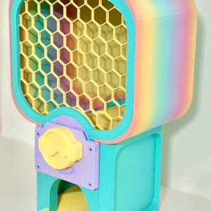 Op de afbeelding: Een kleurrijke kauwgomballenautomaat met een regenboogverloop op de boven- en zijkanten. De machine heeft een geel honingraatraam, een turquoise basis en een gele uitgiftelade. De bedieningsknop is geel en de accenten zijn paars.