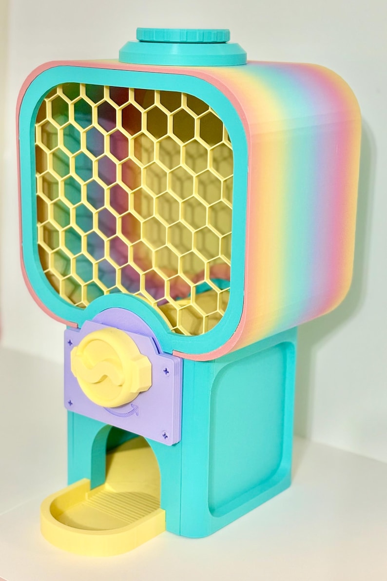 Op de afbeelding: Een kleurrijke kauwgomballenautomaat met een regenboogverloop op de boven- en zijkanten. De machine heeft een geel honingraatraam, een turquoise basis en een gele uitgiftelade. De bedieningsknop is geel en de accenten zijn paars.
