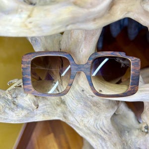 Può includere: Occhiali da sole quadrati con un motivo a venature di legno scuro. Le lenti sono di colore marrone sfumato. Gli occhiali da sole sono appoggiati su una superficie di legno chiaro e strutturato.