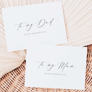 May include: Two white cards with black calligraphy text that reads "To my Dad ON MY WEDDING DAY" and "To my Mum ON MY WEDDING DAY". The cards are on a woven surface.