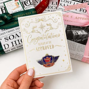 May include: A white greeting card with gold foil accents featuring a bow, stars, and reindeer illustrations. The text reads Congratulations Member of the Approved. A metal enamel pin shaped like a winged shield with a red sleigh and the text Santa's Sleigh Flight Crew is attached to the centre of the card.