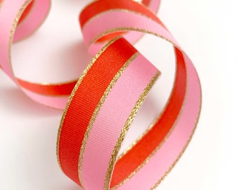 May include: A close-up of a decorative ribbon with a repeating pattern of red and pink stripes. The ribbon has gold trim and is curled in a spiral shape against a white background.