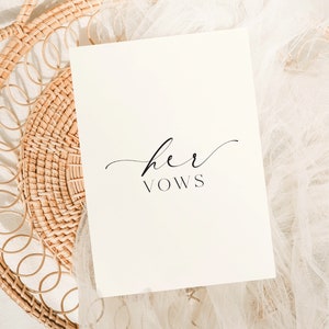 May include: A white card with the text "her vows" written in black cursive script. The card is lying on a white fabric background.