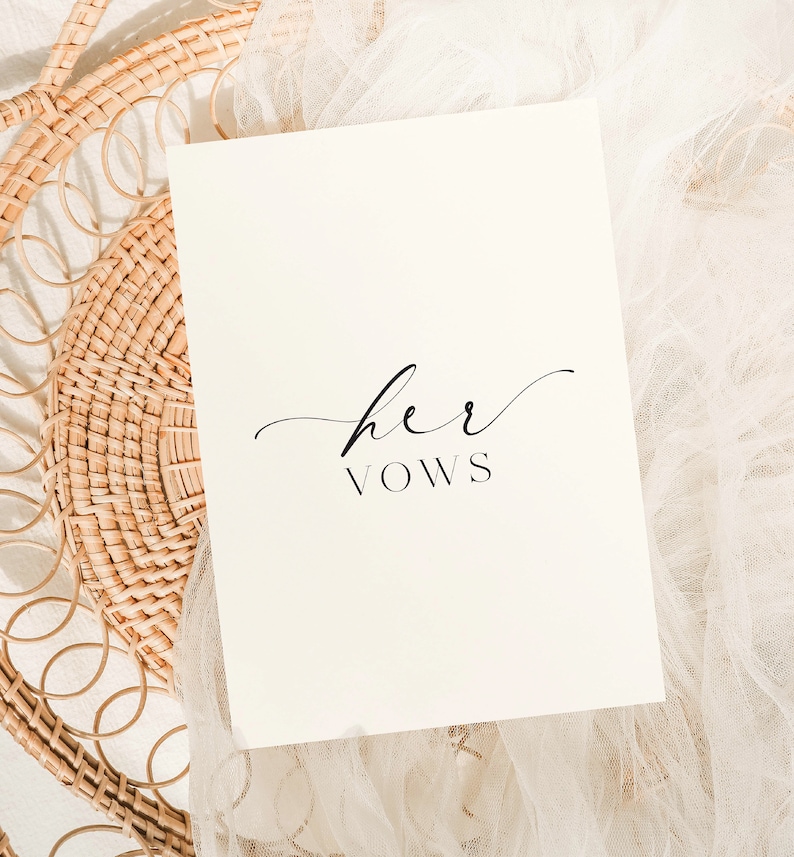 May include: A white card with the text "her vows" written in black cursive script. The card is lying on a white fabric background.