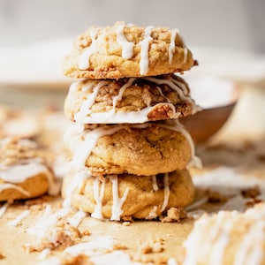 May include: A stack of four cookies with white icing drizzled on top. The cookies are on a brown surface with crumbs scattered around them.