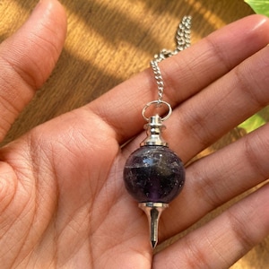 May include: A spherical purple amethyst gemstone pendulum featuring a polished silver-toned metal cap and a pointed silver-toned metal base. The pendulum is attached to a delicate silver-toned metal chain link, creating a balanced, symmetrical design.