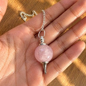 Puede incluir: Un péndulo con una piedra de cuarzo rosa esférica de color rosa pálido, situada entre una tapa de metal plateado y una punta de metal plateado. La piedra está unida a una delicada cadena de metal plateado para su suspensión y movimiento.