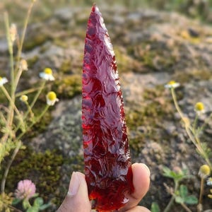 May include: A translucent, deep red, pointed stone blade featuring intricate, hand-knapped flaking patterns along its entire surface. The object is shaped like a spear point with a sharp tip and a slightly wider, notched base, displaying a glass-like texture.