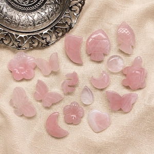 Peut inclure: Une collection de petites sculptures en quartz rose poli aux formes variées, incluant des papillons, des fleurs, des lunes, une tortue et une goutte, disposées près d'un bol décoratif en métal argenté au motif filigrane complexe.