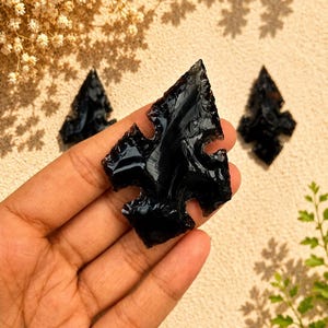 May include: A glossy, black obsidian arrowhead with a sharp, pointed tip and notched sides. The stone features a smooth, reflective surface with distinct flaked edges, showcasing a dark, volcanic glass texture in a triangular, symmetrical shape.