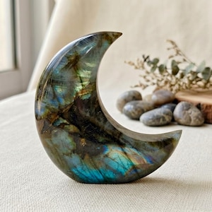 May include: A polished labradorite stone carved into a crescent moon shape. The stone features a dark base with iridescent flashes of blue, green, and gold throughout its surface, displaying natural mineral inclusions and a smooth, reflective finish.