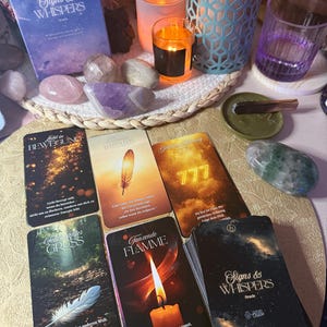 May include: An oracle card deck titled "Signs & Whispers" with several cards fanned out. The cards feature images of feathers, flames, and numbers. The deck is accompanied by crystals, candles, and a small dish with a cinnamon stick.