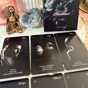 May include: A black and white Lenormand oracle card deck with several cards displayed. The cards feature illustrations and German text. The box has a floral design and the title "The Black Lenormand". A gold figurine and crystals are also visible.