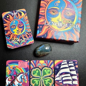 May include: A colorful Crazy Lenormand tarot card deck with a box featuring a sun and moon design. Three cards are displayed, including a rider on a unicorn, a four-leaf clover, and a ship. A polished stone is also present.