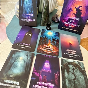 May include: A deck of oracle cards with the title "Shadows of the Ghost Night" in German. The cards feature illustrations of a forest, a clock tower, a bonfire, and a tombstone. The box and cards have a purple and blue color scheme.