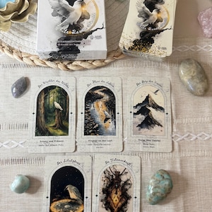 May include: A deck of "White Raven Oracle" cards with a white dove illustration, alongside several individual cards featuring nature-themed artwork. The cards are arranged on a woven tray with decorative stones.