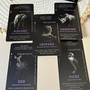 May include: A deck of black tarot cards featuring animal illustrations and German text. The cards depict an elephant, rhinoceros, leopard, deer, and falcon. The deck is titled "Spirit Animal Black Edition."