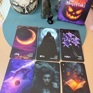 May include: A collection of tarot cards with gothic illustrations, including a card with a woman holding a lantern and another with an open book. The cards are arranged with a decorative plate and a crystal. The box reads "Spooky Leno Rmand".