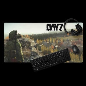 Puede incluir: Una alfombrilla de ratón rectangular para juegos con una escena de paisaje y la palabra "DAYZ" en negro. Un teclado, un ratón y unos auriculares negros están colocados sobre la alfombrilla. La escena representa una figura con máscara de gas.