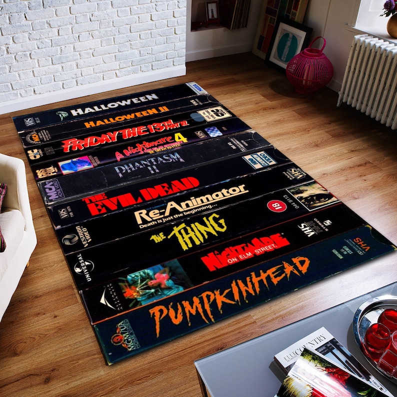 May include: A black and red rug with a design of VHS tapes featuring horror movie titles like "Halloween", "Friday the 13th", "A Nightmare 4", "Phantasm", "Evil Dead", "Re-Animator", "The Thing", "Nightmare on Elm Street", and "Pumpkinhead".