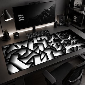May include: Black and white computer desk setup featuring a large monitor displaying a mountain scene and the words "FOCUS CREATE INSPIRE". A geometric patterned mousepad dominates the desk, alongside a keyboard, speakers, and a desk lamp.