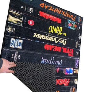 May include: A black and white rug with a design of VHS tapes featuring horror movie titles like "Phantasm", "Evil Dead", "Re-Animator", "The Thing", "Nightmare on Elm Street", and "Pumpkinhead".