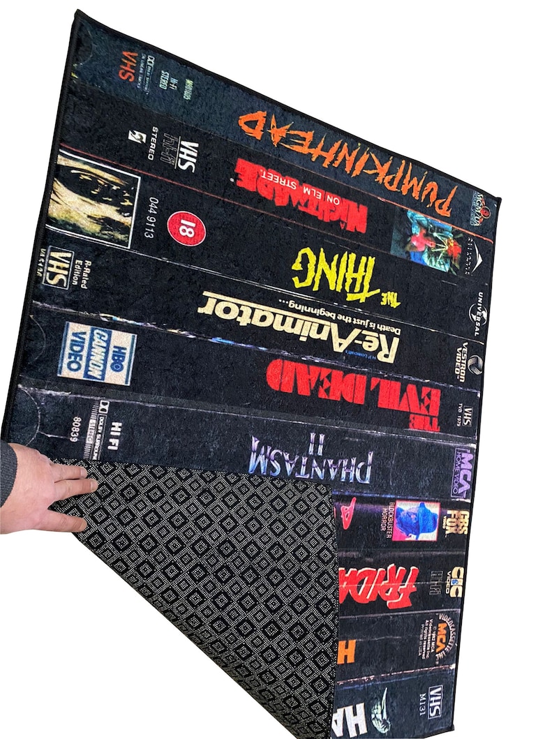 May include: A black and white rug with a design of VHS tapes featuring horror movie titles like "Phantasm", "Evil Dead", "Re-Animator", "The Thing", "Nightmare on Elm Street", and "Pumpkinhead".