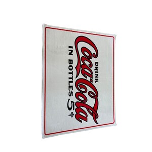 K&ouml;nnte beinhalten: Ein wei&szlig;es rechteckiges Schild mit einem roten Coca-Cola-Logo und dem Text "Drink Coca-Cola in bottles 5&cent;".