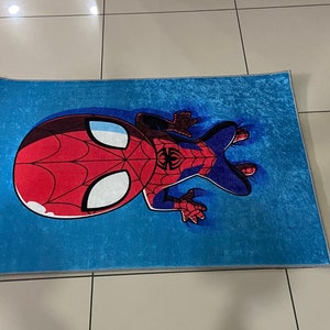 May include: A blue rug with a cartoon image of Spiderman in a red and blue suit. The rug is made of a soft, plush material.
