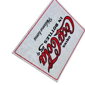 K&ouml;nnte beinhalten: Ein wei&szlig;es Schild mit rotem Text, auf dem "Drink Coca-Cola in Bottles" und "Welcome Home" steht.