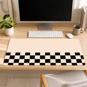 Op de afbeelding: Een beige rechthoekige bureaumat met een zwart-wit geblokt patroon langs de onderrand. De mat ligt op een houten bureau, met een computermonitor, toetsenbord, muis en een mok zichtbaar.