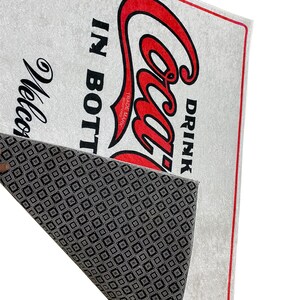 K&ouml;nnte beinhalten: Eine wei&szlig;e Fu&szlig;matte mit rotem Rand und einem roten Coca-Cola-Logo mit dem Text "Drink Coca-Cola in Bottles" und dem Markenzeichen. Die R&uuml;ckseite der Fu&szlig;matte hat ein schwarz-wei&szlig;es Rautenmuster.