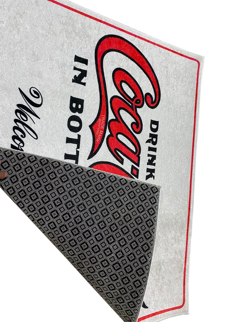 K&ouml;nnte beinhalten: Eine wei&szlig;e Fu&szlig;matte mit rotem Rand und einem roten Coca-Cola-Logo mit dem Text "Drink Coca-Cola in Bottles" und dem Markenzeichen. Die R&uuml;ckseite der Fu&szlig;matte hat ein schwarz-wei&szlig;es Rautenmuster.