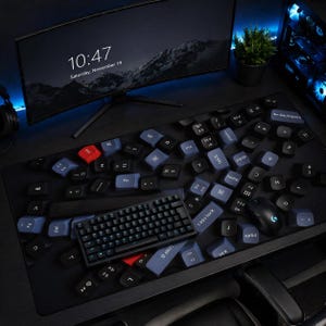 May include: A black desk setup featuring a computer monitor displaying the time and date. A black keyboard, mouse, and scattered keycaps are on a large black mousepad. The keycaps are blue, black, and red.