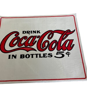 K&ouml;nnte beinhalten: Ein wei&szlig;es rechteckiges Schild mit dem roten und schwarzen Coca-Cola-Logo und dem Text "Drink Coca-Cola in Bottles 5&cent;".