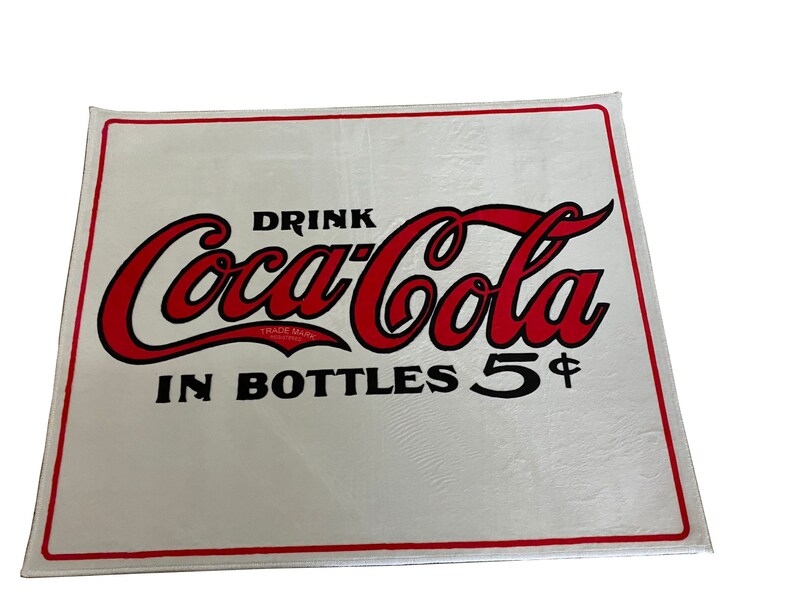 K&ouml;nnte beinhalten: Ein wei&szlig;es rechteckiges Schild mit dem roten und schwarzen Coca-Cola-Logo und dem Text "Drink Coca-Cola in Bottles 5&cent;".