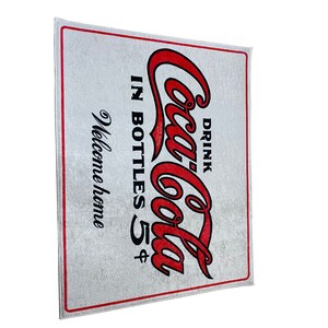 K&ouml;nnte beinhalten: Eine wei&szlig;e Fu&szlig;matte mit einem roten Coca-Cola-Logo und dem Text "Drink Coca-Cola in bottles 5&cent;" und "Welcome home".