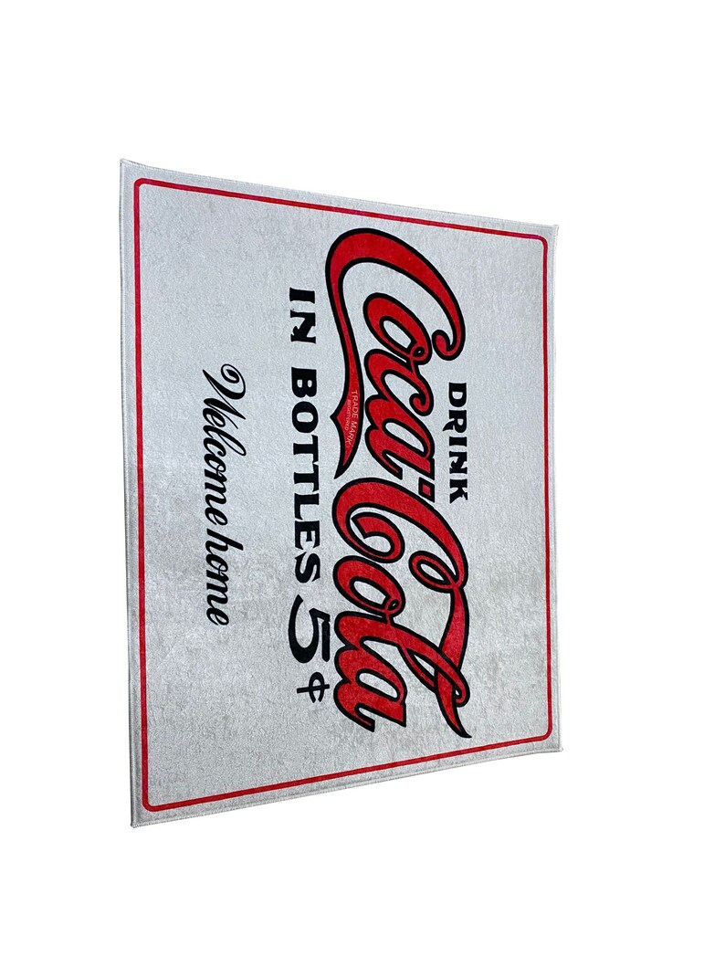 K&ouml;nnte beinhalten: Eine wei&szlig;e Fu&szlig;matte mit einem roten Coca-Cola-Logo und dem Text "Drink Coca-Cola in bottles 5&cent;" und "Welcome home".