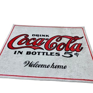 K&ouml;nnte beinhalten: Wei&szlig;e Willkommensmatte mit rotem Coca-Cola-Logo und dem Text "Drink Coca-Cola in Bottles 5&cent; Welcome home".