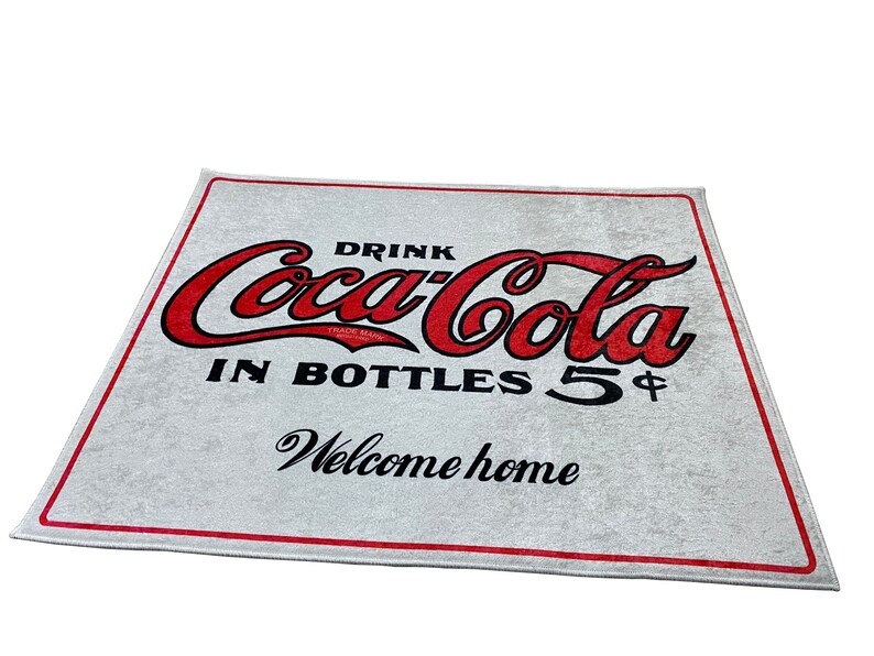K&ouml;nnte beinhalten: Wei&szlig;e Willkommensmatte mit rotem Coca-Cola-Logo und dem Text "Drink Coca-Cola in Bottles 5&cent; Welcome home".