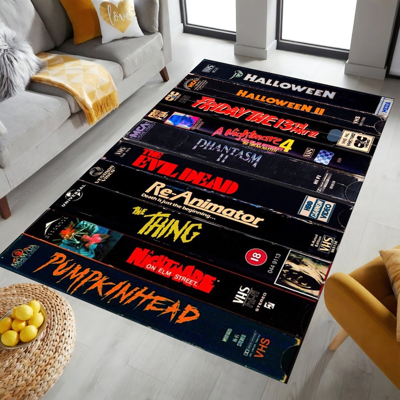 May include: A black and white rug with a design of VHS tapes featuring horror movie titles like "Halloween", "Friday the 13th", "A Nightmare on Elm Street", "Evil Dead", "Re-Animator", "The Thing", and "Pumpkinhead".