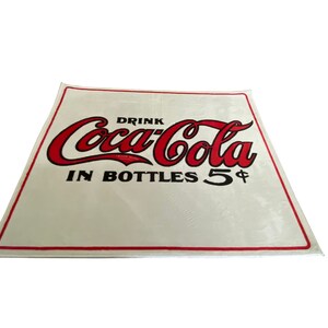 K&ouml;nnte beinhalten: Wei&szlig;es Schild mit rotem Coca-Cola-Logo und schwarzem Text "Drink Coca-Cola in Bottles 5&cent;".