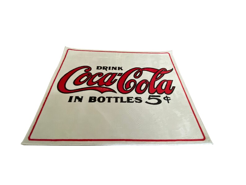 K&ouml;nnte beinhalten: Wei&szlig;es Schild mit rotem Coca-Cola-Logo und schwarzem Text "Drink Coca-Cola in Bottles 5&cent;".