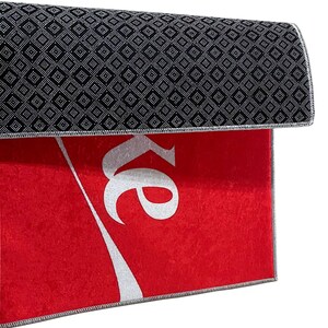 May include: Red doormat with a white Coca-Cola logo. The back of the doormat has a black diamond pattern.