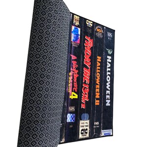 May include: A stack of VHS tapes with the titles "Halloween", "Halloween II", "Friday the 13th", and "A Nightmare on Elm Street 4" on a black and white patterned surface.